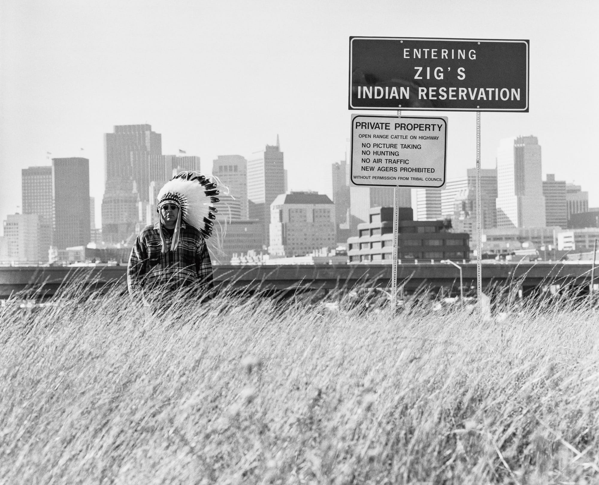 a black-and-white photo documenting an artwork by Zig Jackson, with a sign reading "Entering Zig's Indian Reservation" and a man standing in a Native American feathered chief's war bonnet