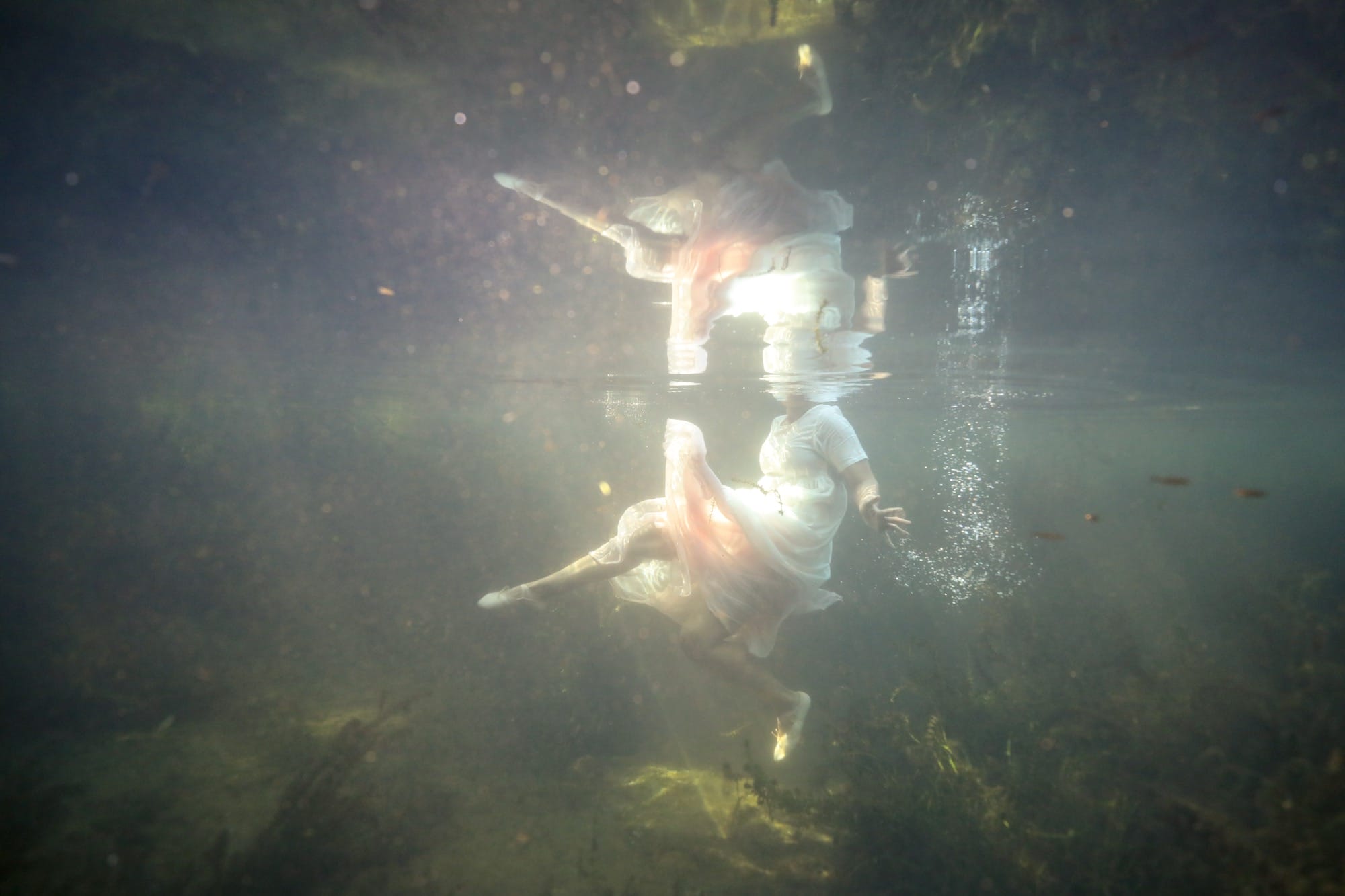a photograph of a figure underwater with the sun shining on their body, head invisible above the water and amid a reflection