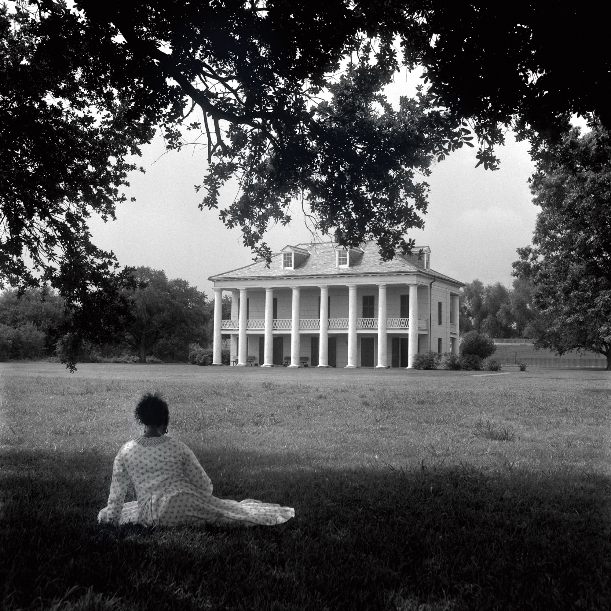a black-and-white photograph by Carrie Mae Weems of a Black woman in a white dress looking at a plantation house